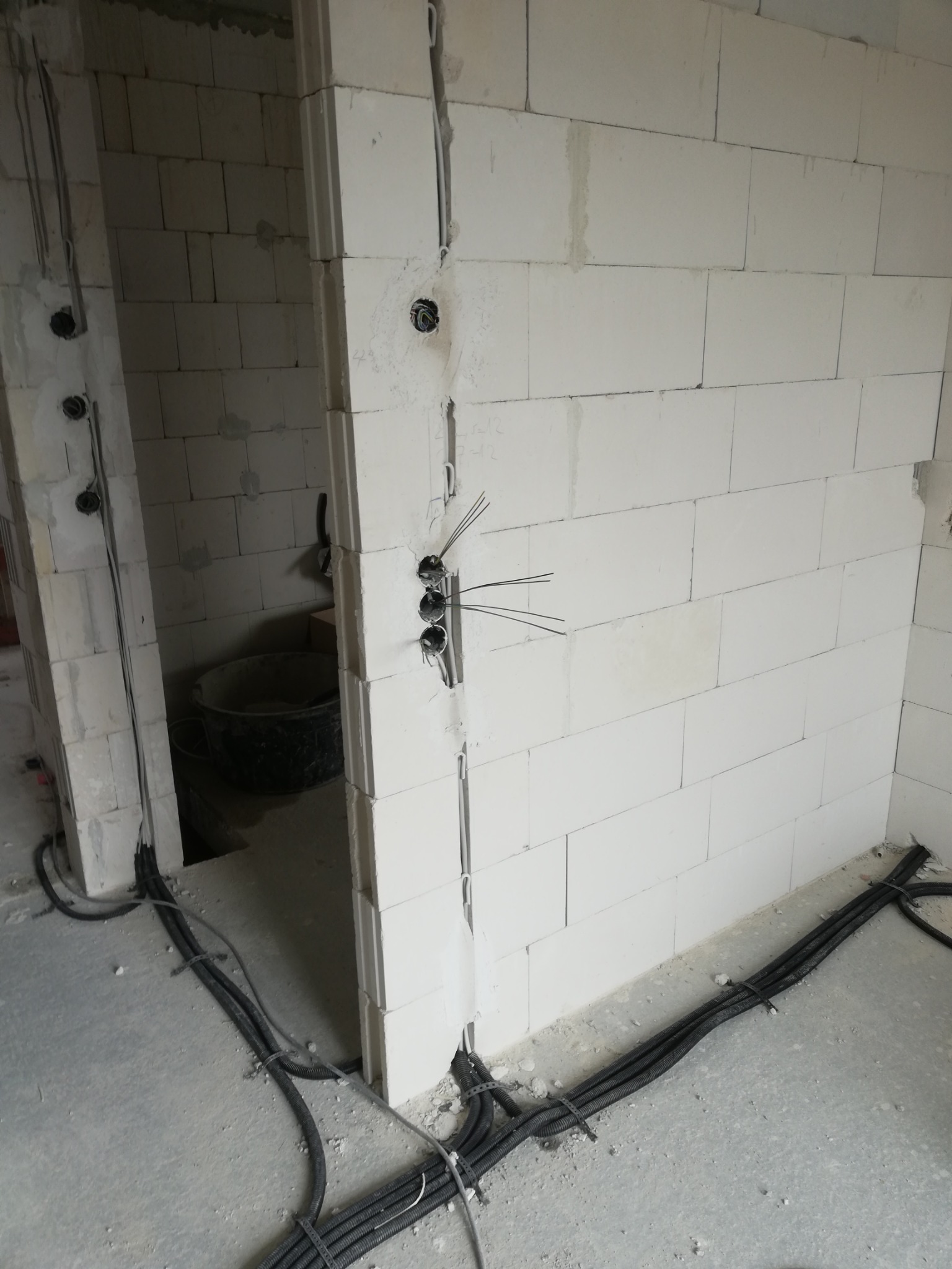 Surowe ściany z betonu komórkowego w trakcie prac instalacyjnych; widoczne przewody elektryczne wystające z puszek w ścianie oraz prowadzone w peszlach po podłodze.