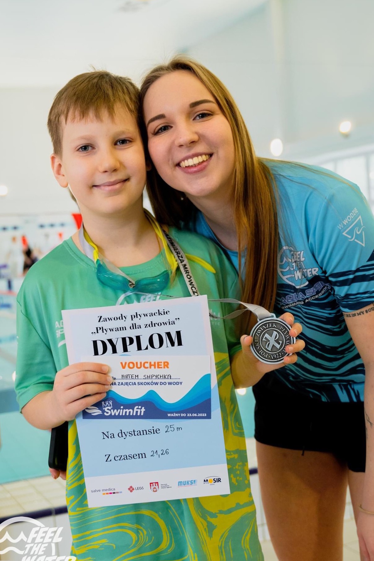 Chłopiec z dyplomem za zawody pływackie 'Pływam dla zdrowia' i voucher na zajęcia skoków do wody, pozuje z uśmiechniętą instruktorką trzymającą medal Związku Pływackiego na tle basenu.