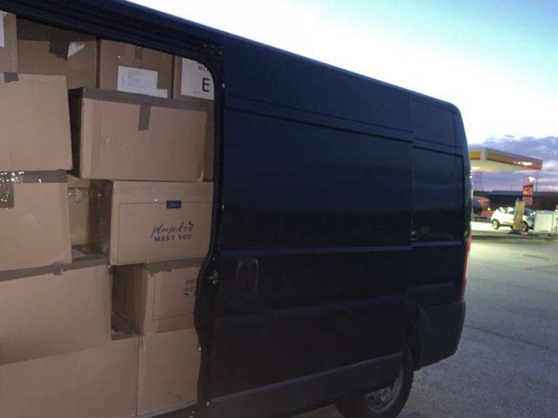 Ciemny van załadowany kartonami z napisem 'Please to MEET YOU' stoi na stacji benzynowej o zmierzchu.