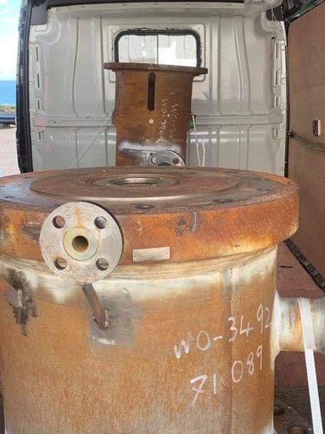 Rdzawe, przemysłowe elementy metalowe z numerami seryjnymi, załadowane do białego furgonetki z otwartymi drzwiami, widoczne morze w tle.