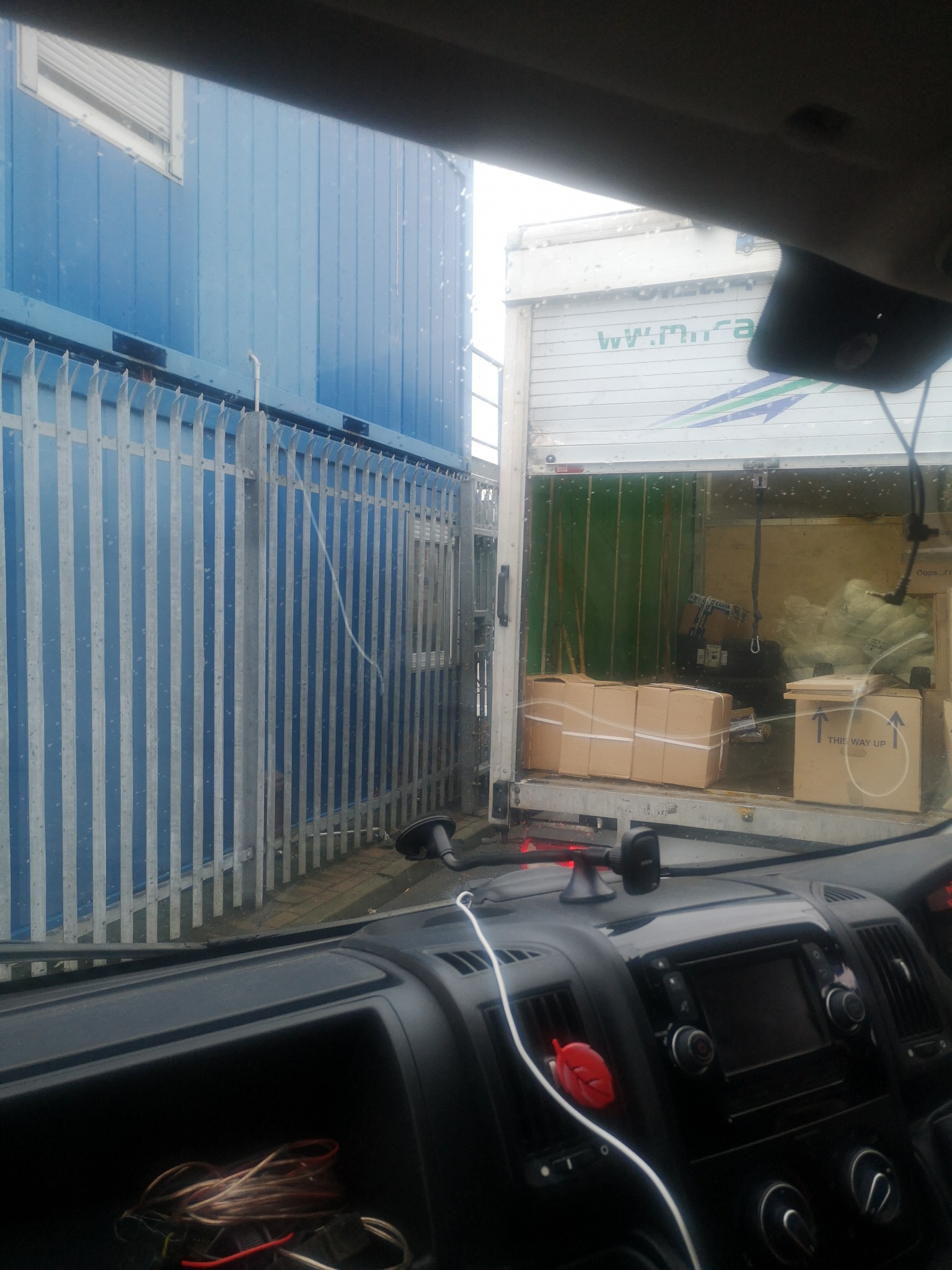 Widok z kabiny samochodu na otwartą pakę ciężarówki załadowną kartonami, obok niebieski kontener biurowy z metalowym ogrodzeniem.