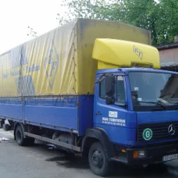 Niebieska ciężarówka Mercedes z żółtą plandeką i nadbudówką, na której widnieje logo firmy transportowej. Na plandece również widoczny znak firmy. Pojazd zaparkowany na asfaltowym placu.