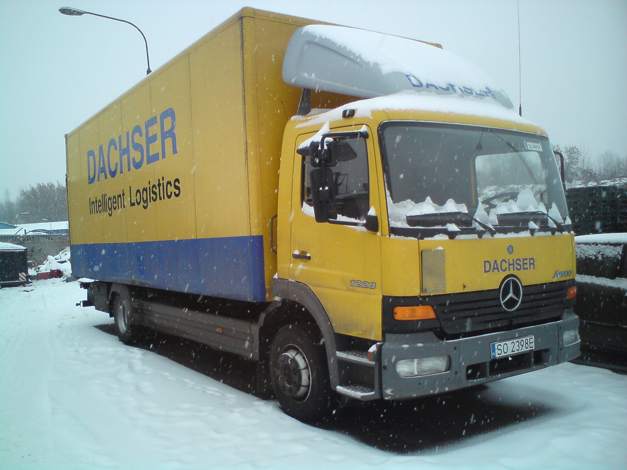 Żółto-niebieska ciężarówka marki Mercedes-Benz Atego z napisem DACHSER, częściowo pokryta śniegiem, zaparkowana na śnieżnym placu w pochmurny dzień.