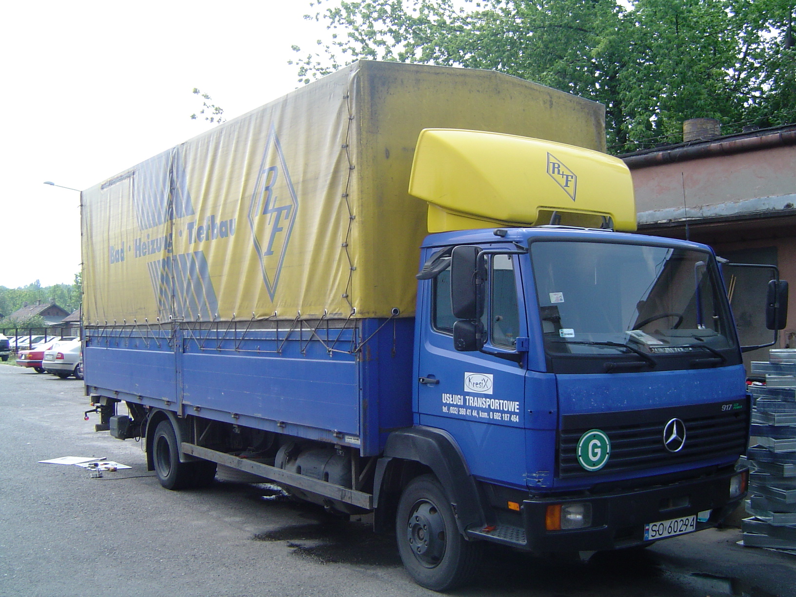 Niebieska ciężarówka Mercedes z żółtą plandeką i nadbudówką, na której widnieje logo firmy transportowej. Na plandece również widoczny znak firmy. Pojazd zaparkowany na asfaltowym placu.