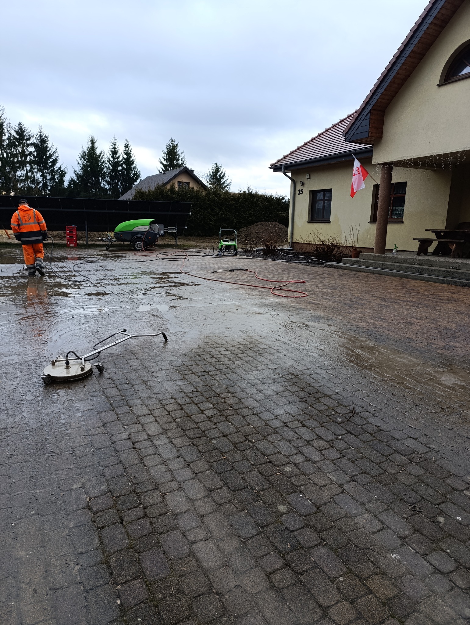 Pracownik w kombinezonie czyści kostkę brukową na posesji domu jednorodzinnego za pomocą myjki ciśnieniowej. Widoczny efekt czyszczenia.