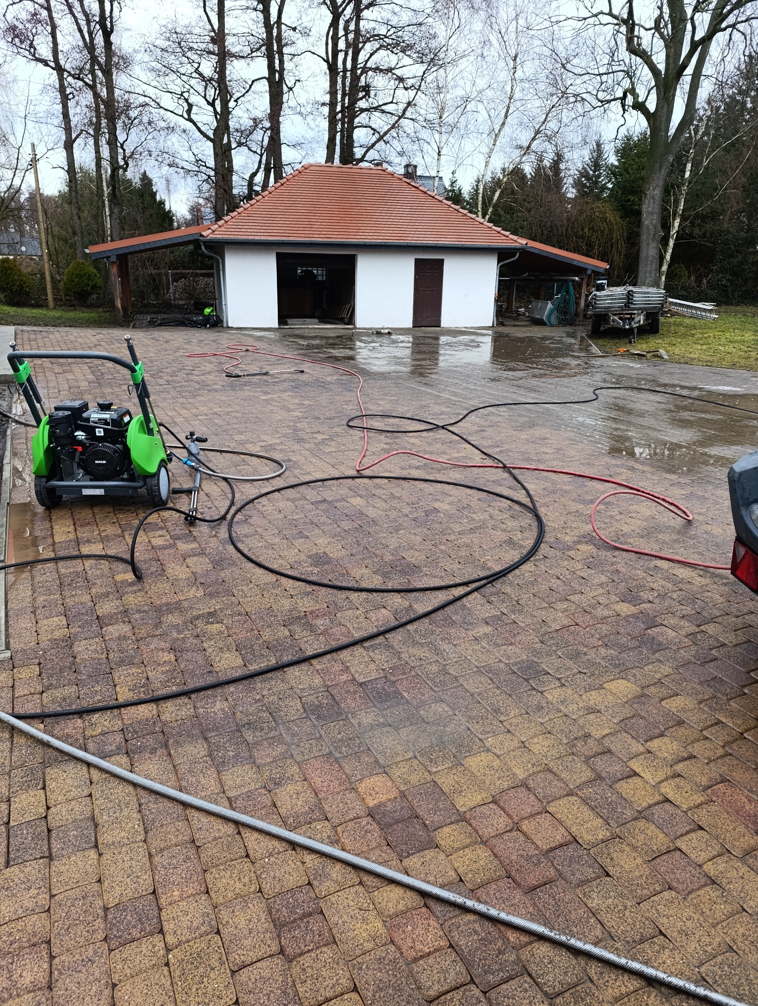 Zielona myjka ciśnieniowa na mokrej kostce brukowej przed garażem z czerwoną dachówką. Węże rozłożone na kostce. Widok na budynek i drzewa w tle.