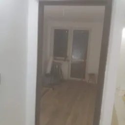 Widok przez ościeżnicę na pokój w trakcie prac wykończeniowych: pomalowane ściany, parkiet na podłodze, grzejnik, okno balkonowe i narzędzia.