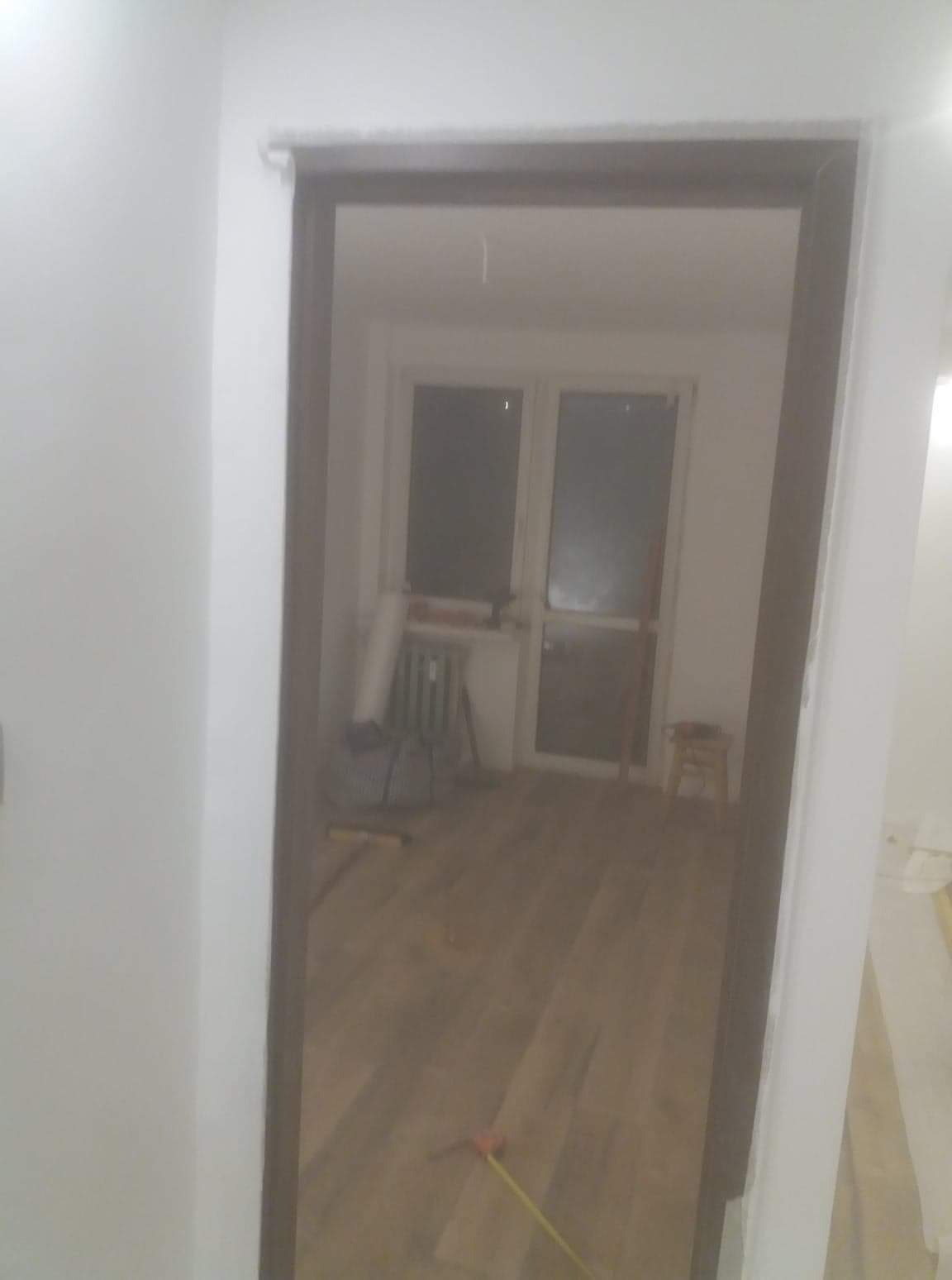 Widok przez ościeżnicę na pokój w trakcie prac wykończeniowych: pomalowane ściany, parkiet na podłodze, grzejnik, okno balkonowe i narzędzia.