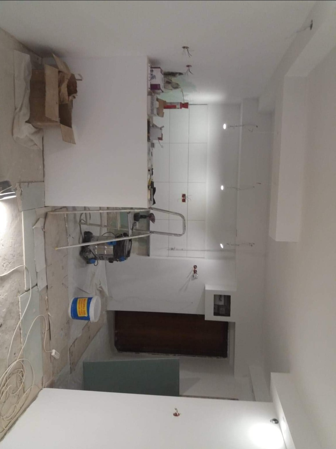 Wnętrze w trakcie remontu: widoczna drabina, agregat malarski, puszka z farbą i płyta karton gips, ściany częściowo przygotowane do malowania, sufit z punktowym oświetleniem w trakcie instalacji.