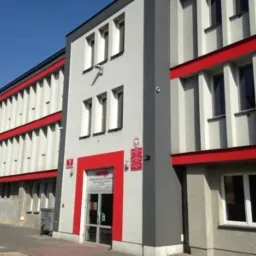 Fasada budynku szkoleniowego z szarymi ścianami i czerwonymi akcentami, widoczne okna i wejście z logo firmy szkoleniowej.