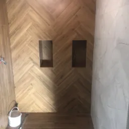 Ukończona łazienka z drewnopodobnymi płytkami ułożonymi w jodełkę na ścianie z wnękami, na podłodze odpływ liniowy, ściana wyłożona marmurowymi płytkami, widoczne wiadro po farbie.