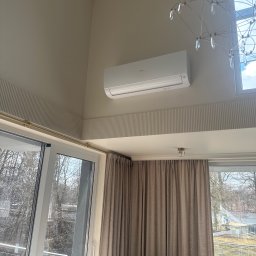 CLIMETIQA SPÓŁKA Z OGRANICZONĄ ODPOWIEDZIALNOŚCIĄ - Biały klimatyzator Haier zamontowany na ścianie w jasnym wnętrzu z dużym oknem i dekoracyjną listwą. Widoczny fragment żyrandola.