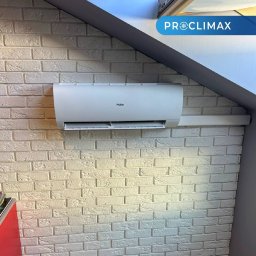 CLIMETIQA SPÓŁKA Z OGRANICZONĄ ODPOWIEDZIALNOŚCIĄ - Biały klimatyzator Haier zamontowany na ścianie z dekoracyjnej cegły w pomieszczeniu ze skośnym sufitem. Widoczny fragment logo firmy Proclimax.