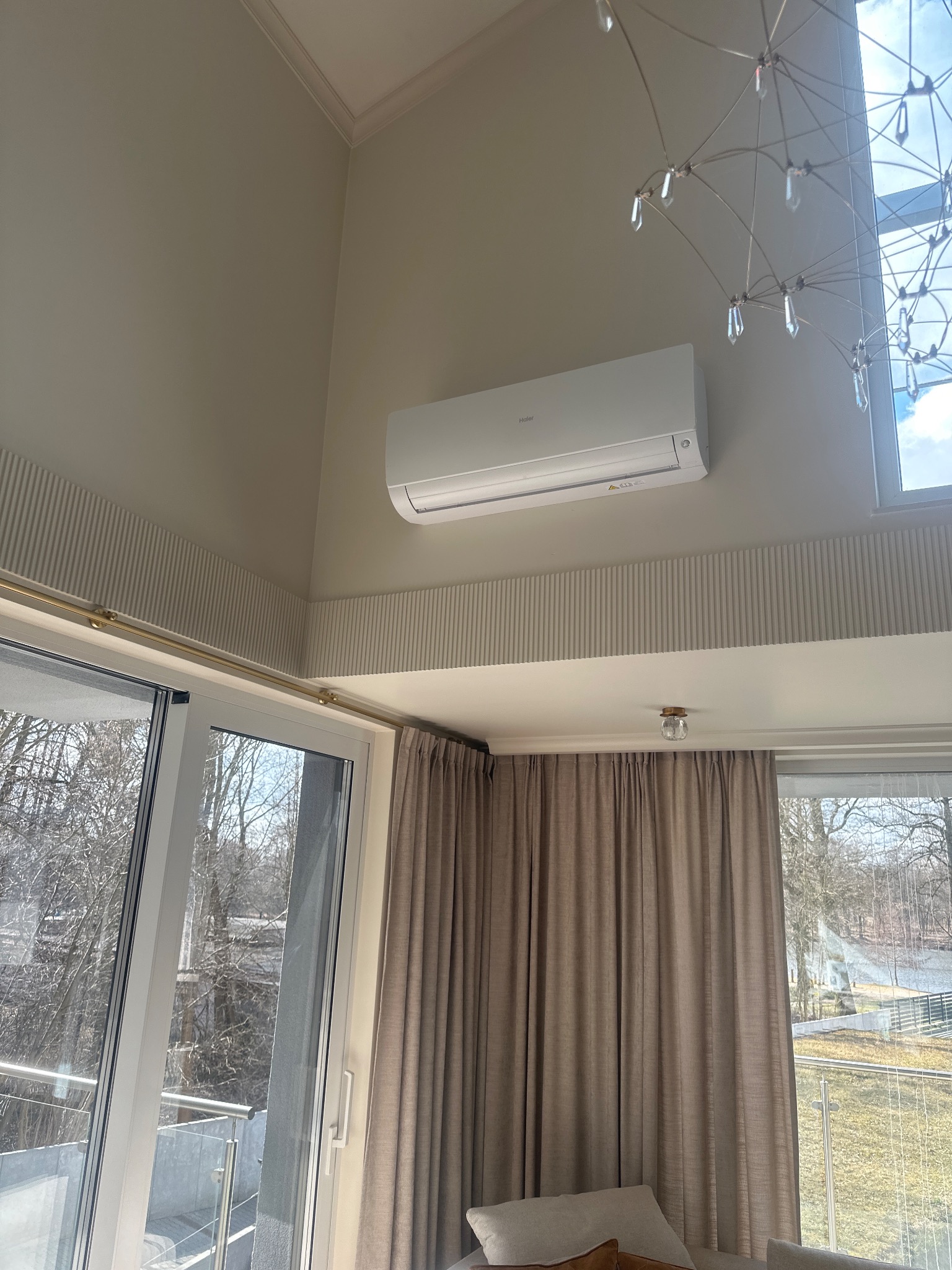 Biały klimatyzator Haier zamontowany na ścianie w jasnym wnętrzu z dużym oknem i dekoracyjną listwą. Widoczny fragment żyrandola.