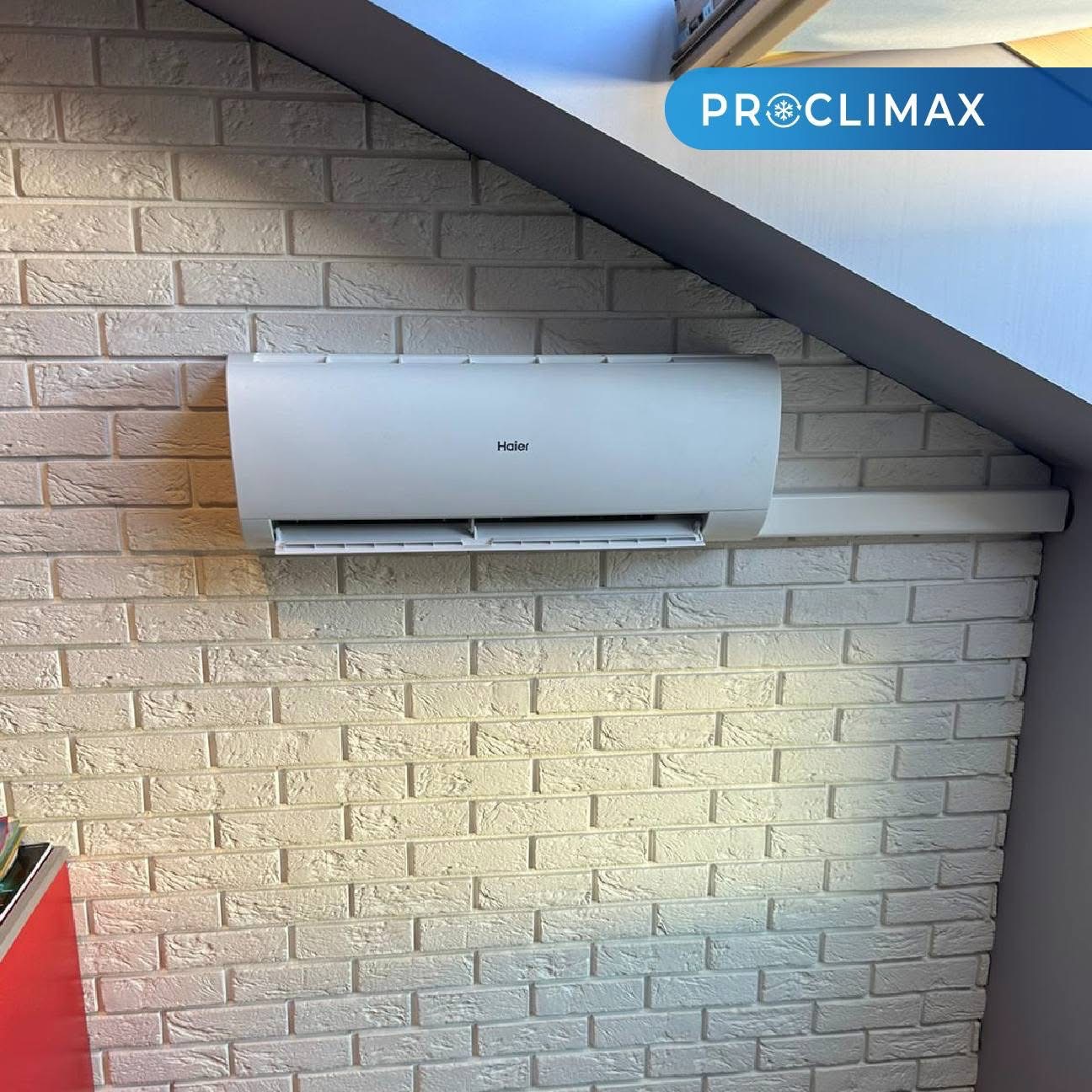 Biały klimatyzator Haier zamontowany na ścianie z dekoracyjnej cegły w pomieszczeniu ze skośnym sufitem. Widoczny fragment logo firmy Proclimax.