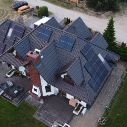 Widok z drona na dom z ciemną dachówką, na której zamontowano panele słoneczne. Widoczne również dwa samochody na podjeździe i ogród z meblami.