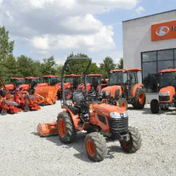 Pomarańczowe traktory Kubota B1241 i inny sprzęt rolniczy na żwirowym placu przed salonem firmowym Kubota, widok na budynek z dużymi oknami i logo firmy.