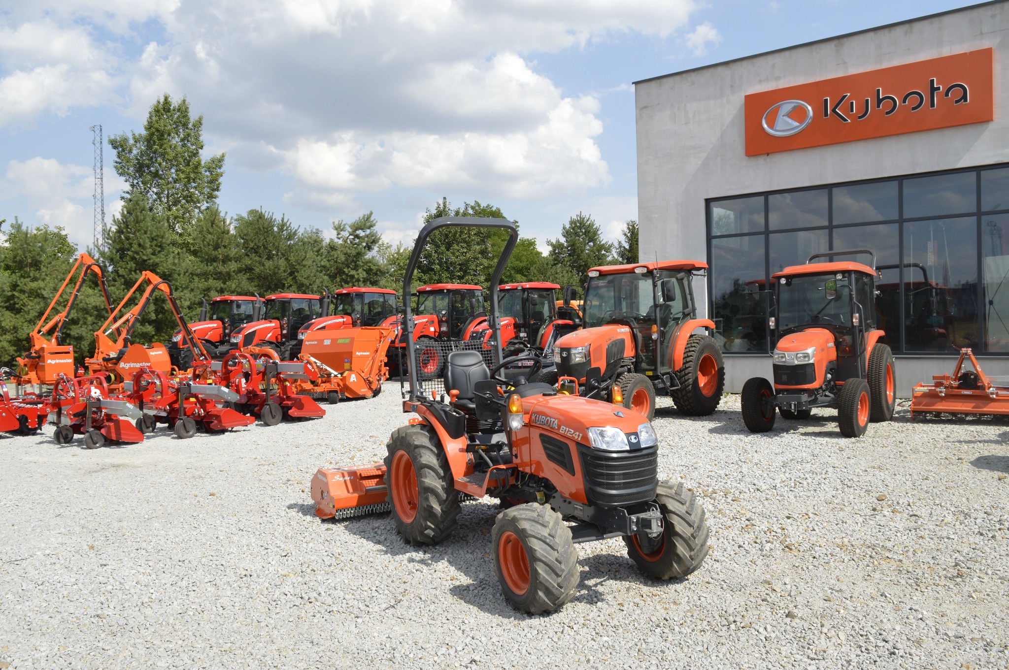 Pomarańczowe traktory Kubota B1241 i inny sprzęt rolniczy na żwirowym placu przed salonem firmowym Kubota, widok na budynek z dużymi oknami i logo firmy.