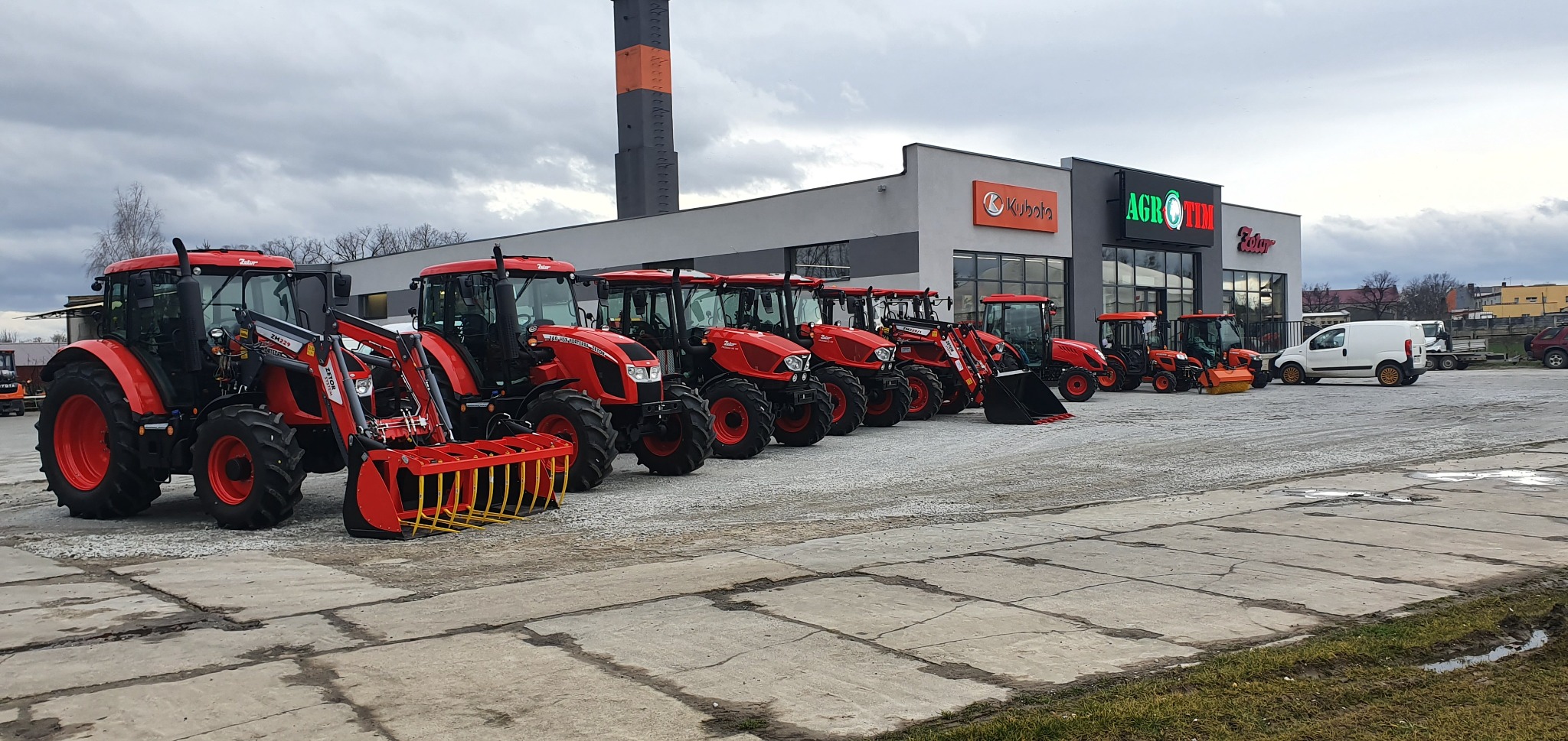 Rząd czerwonych traktorów Zetor i Kubota z różnym osprzętem, stojących przed salonem sprzedaży maszyn rolniczych AgroTim i Kubota w pochmurny dzień.