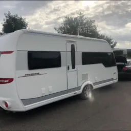 Biała przyczepa kempingowa marki Hobby Premium, zaparkowana na poboczu drogi, z podłączonym czarnym samochodem osobowym z otwartym bagażnikiem.