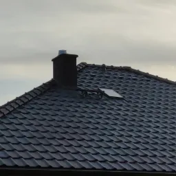 Ciemnoszary dach z dachówki ceramicznej z kominem i oknem dachowym na tle pochmurnego nieba.