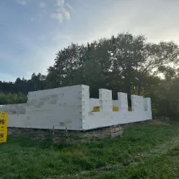 Wznoszenie ścian parterowego domu z białych bloczków Ytong na tle zieleni, widoczne palety z materiałem budowlanym.