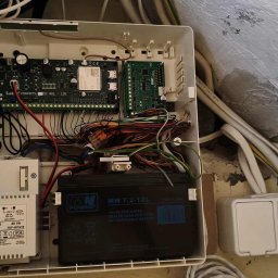 F-VISION Filip Rabęda - Otwarta skrzynka alarmowa z płytą główną, akumulatorem i transformatorem. Widoczne liczne przewody i złącza. Instalacja w trakcie montażu.