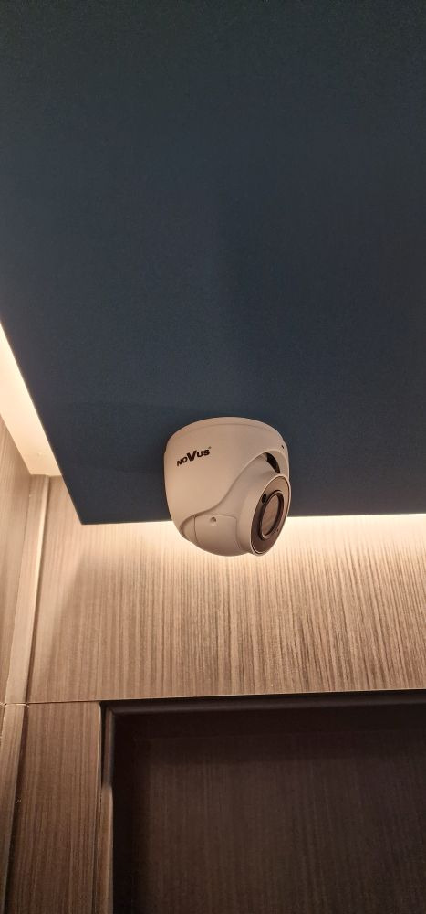 Kamera Novus zamontowana na suficie w pomieszczeniu. Widoczne oświetlenie LED i elementy wykończenia wnętrza. Ujęcie z dołu.