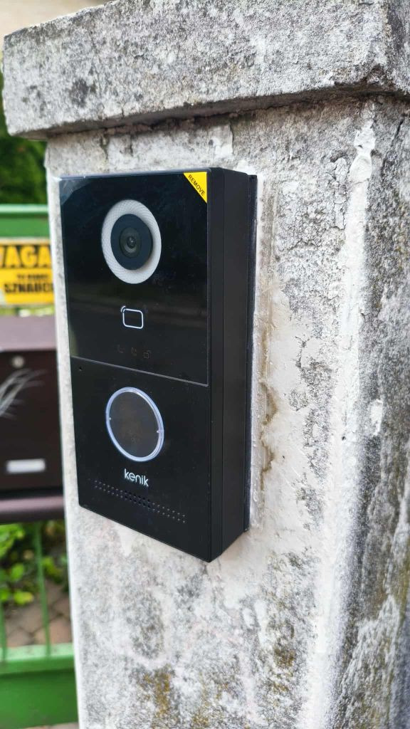 Czarny wideodomofon Kenik z kamerą zamontowany na szarej, betonowej kolumnie we Wrocławiu. Widoczny fragment skrzynki pocztowej i zielonego ogrodzenia.