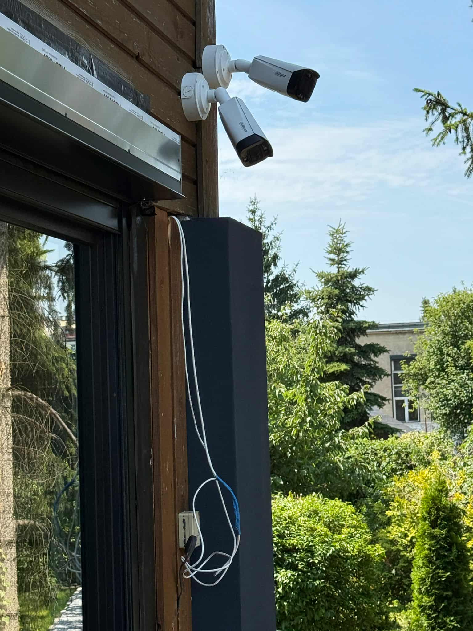 Dwie kamery monitoringu Dahua zamontowane na drewnianej elewacji budynku, z widocznymi kablami i otaczającą zielenią. Widok na błękitne niebo.