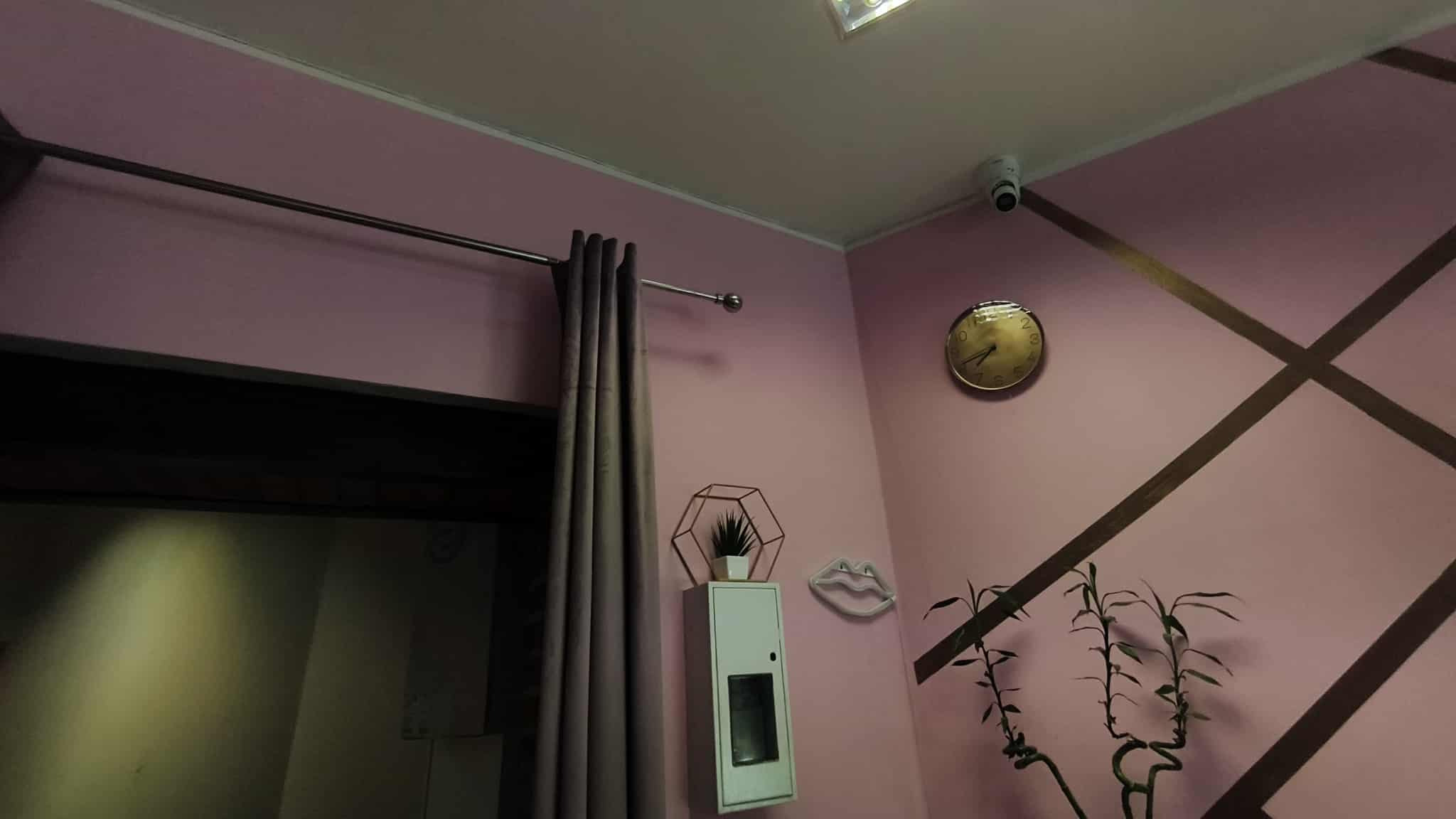 Kamera IP na suficie w pokoju z różowymi ścianami, zegarem i dekoracjami. Widoczna szafka z zamontowanym panelem sterowania. Minimalistyczny design.