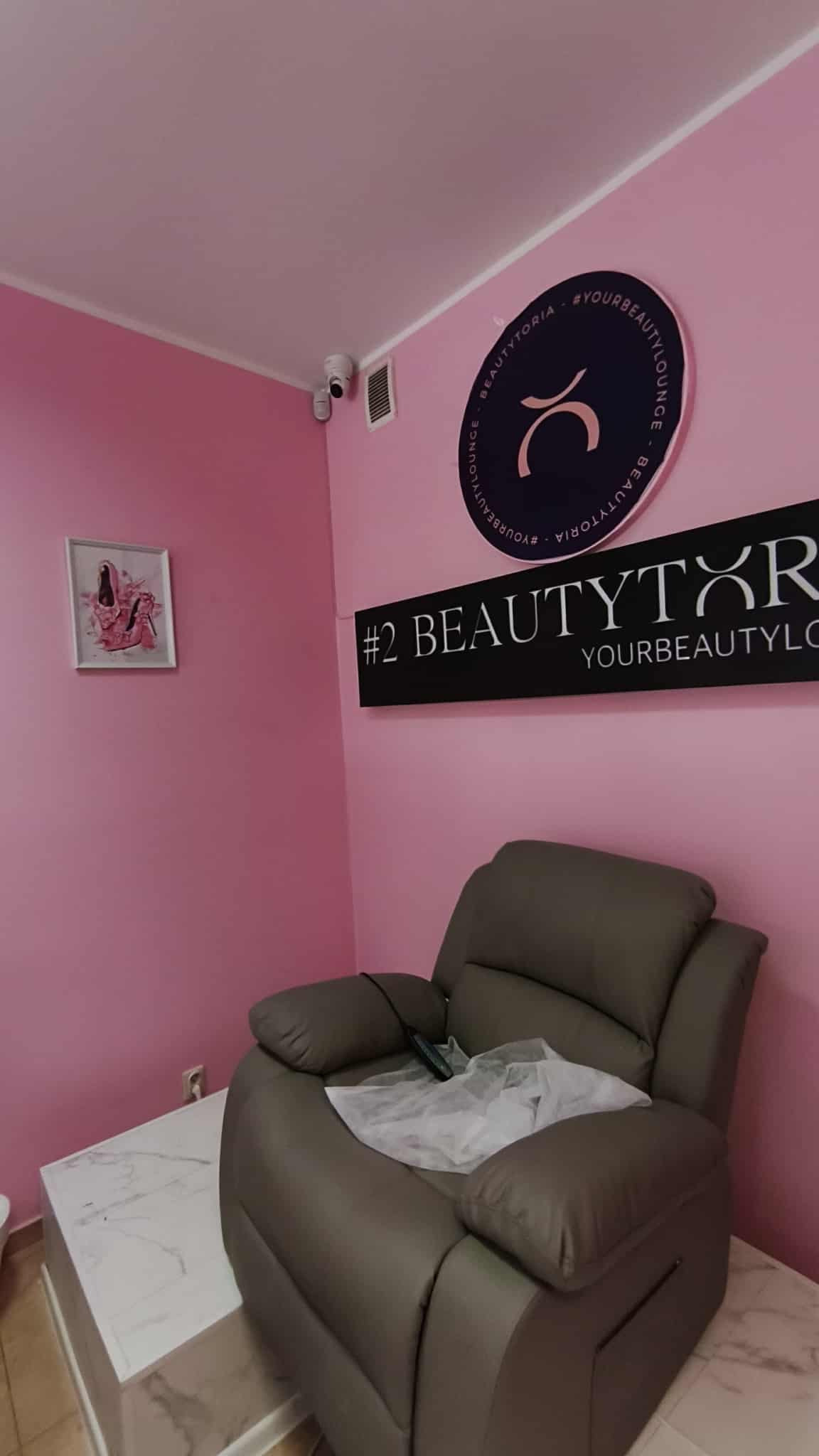 Różowe wnętrze salonu BeautyToria z kamerą monitoringu, logotypami, fotelem relaksacyjnym i obrazkiem baletek. Nowoczesny design i dbałość o bezpieczeństwo.