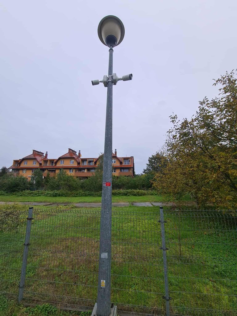 Słup oświetleniowy z dwoma kamerami monitoringu na tle budynku mieszkalnego, widziany z dołu przez ogrodzenie z siatki.