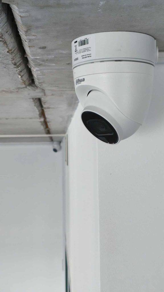 Biała kamera kopułkowa Dahua zamontowana pod betonowym sufitem, widoczna naklejka z kodem kreskowym i logo. Druga kamera w tle.