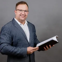 Uśmiechnięty rzeczoznawca w okularach i marynarce trzyma otwarty raport budowlany na szarym tle.