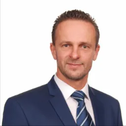 Portret mężczyzny w niebieskim garniturze i krawacie w paski, z lekkim uśmiechem i krótką brodą na białym tle.