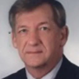 Portret mężczyzny w średnim wieku, w ciemnym garniturze i białej koszuli, z krótkimi, zaczesanymi do tyłu włosami i łagodnym uśmiechem, na rozmytym tle.