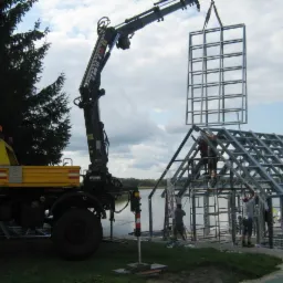 Żółty dźwig samochodowy z wysięgnikiem podnosi stalową ramę budynku na brzegu jeziora, gdzie grupa robotników montuje konstrukcję szkieletową.