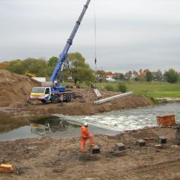 Usługi dzwigowe , Pomoc drogowa TIR , Parking Strzeżony Grzegorz Leymańczyk - Żuraw samochodowy w trakcie montażu betonowych elementów konstrukcyjnych w korycie rzeki, pracownik w pomarańczowym kombinezonie nadzoruje proces, w tle widoczny krajobraz wiejski.