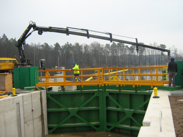 Czarny dźwig samochodowy z wysięgnikiem nad zieloną konstrukcją śluzy wodnej, pracownik w żółtej kurtce na pomostach, w tle zbiornik wodny i drzewa.