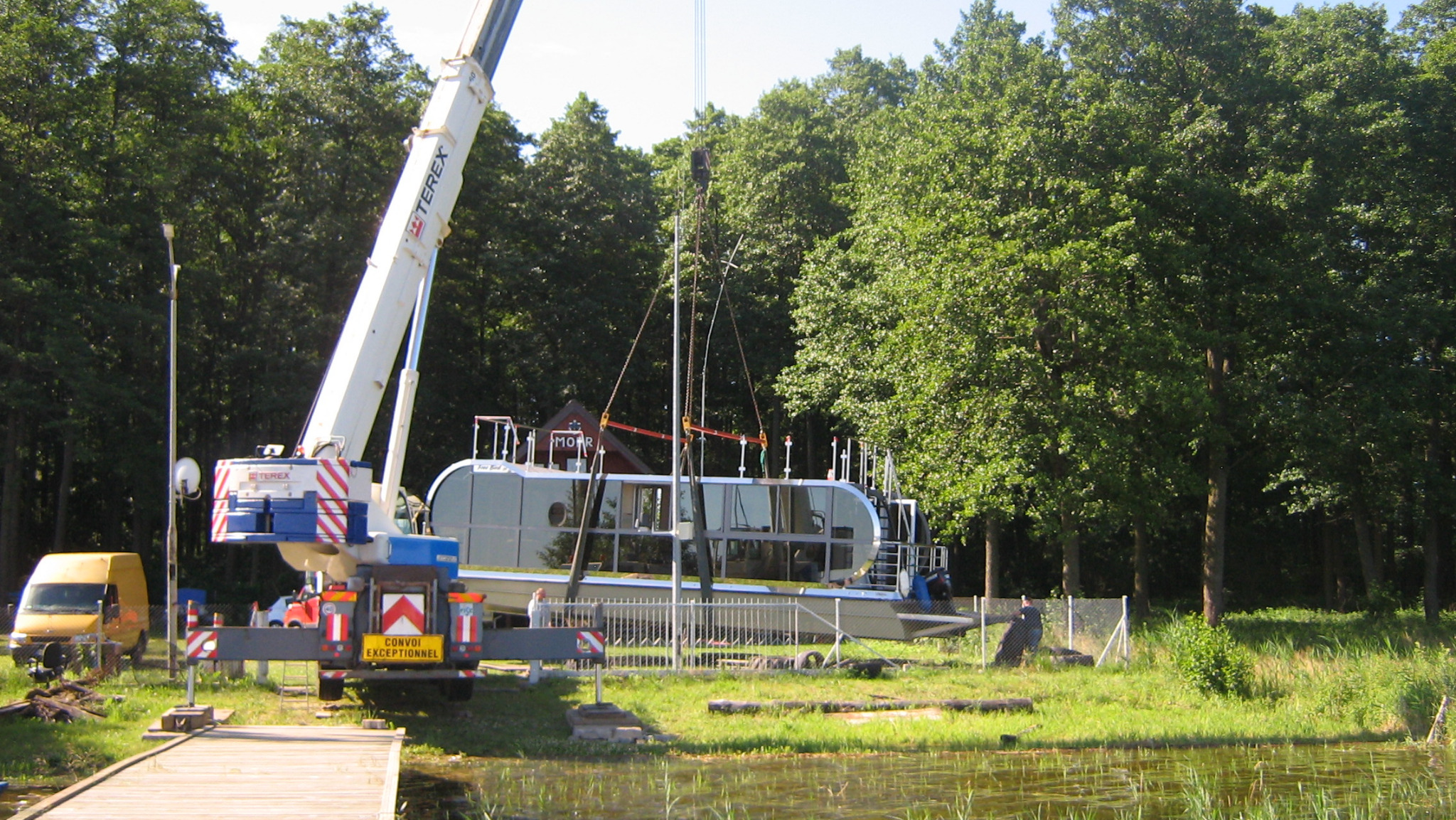 Biały dźwig Terex podnosi przeszkloną łódź z napisem 'MOOR' nad brzegiem jeziora, otoczonego zielonymi drzewami; żółty van w tle.