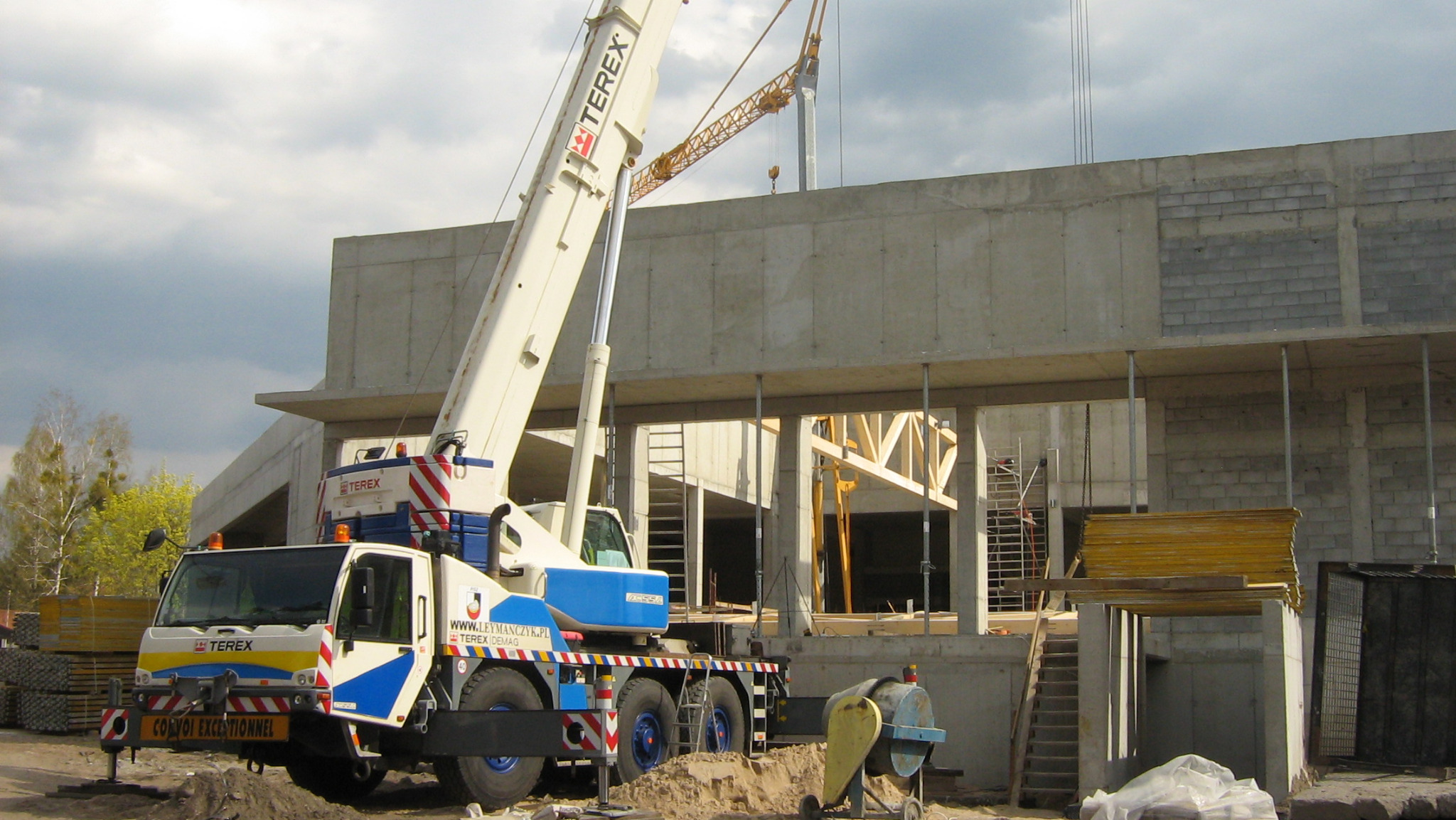 Żuraw samojezdny Terex na placu budowy, w tle surowy budynek z betonu i cegły, widoczna konstrukcja drewniana wewnątrz budynku.