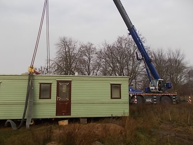 Przenoszenie zielonego baraku mieszkalnego za pomocą niebieskiego dźwigu teleskopowego HHL na tle pochmurnego nieba i drzew bez liści.