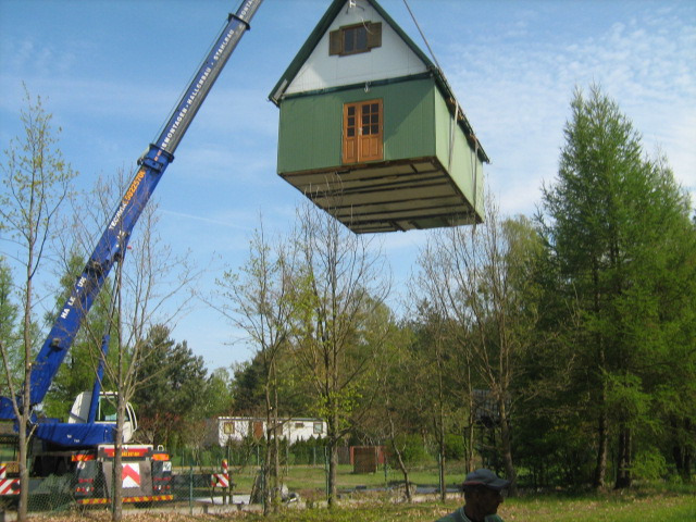 Przenoszenie gotowego, zielonego domku jednorodzinnego za pomocą dźwigu na tle drzew i błękitnego nieba.