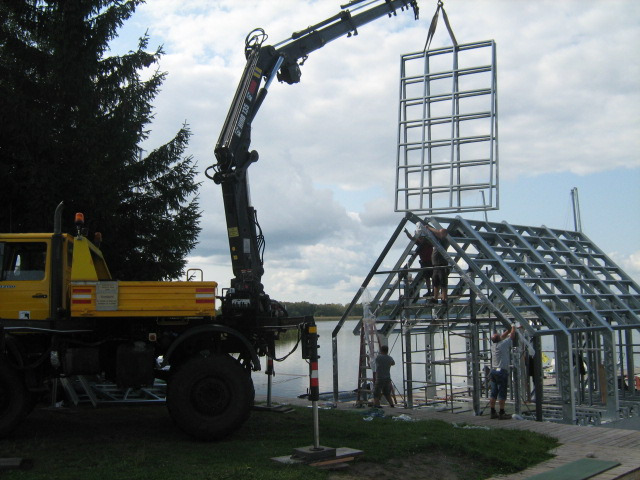 Żółty dźwig samochodowy z wysięgnikiem podnosi stalową ramę budynku na brzegu jeziora, gdzie grupa robotników montuje konstrukcję szkieletową.