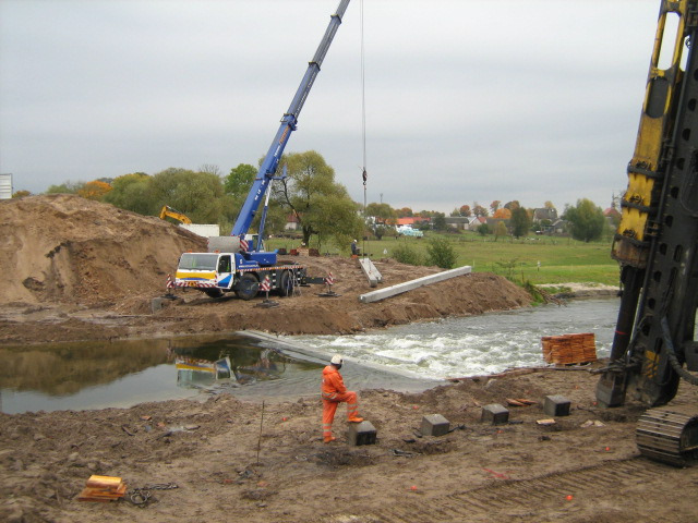 Żuraw samochodowy w trakcie montażu betonowych elementów konstrukcyjnych w korycie rzeki, pracownik w pomarańczowym kombinezonie nadzoruje proces, w tle widoczny krajobraz wiejski.