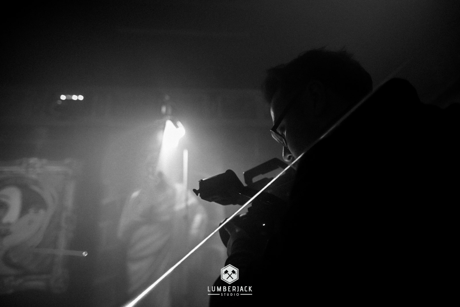 Czarno-białe zdjęcie operatora kamery z profilu, z widocznym laserem pomiarowym, w tle rozmyte postacie i oświetlenie sceniczne.