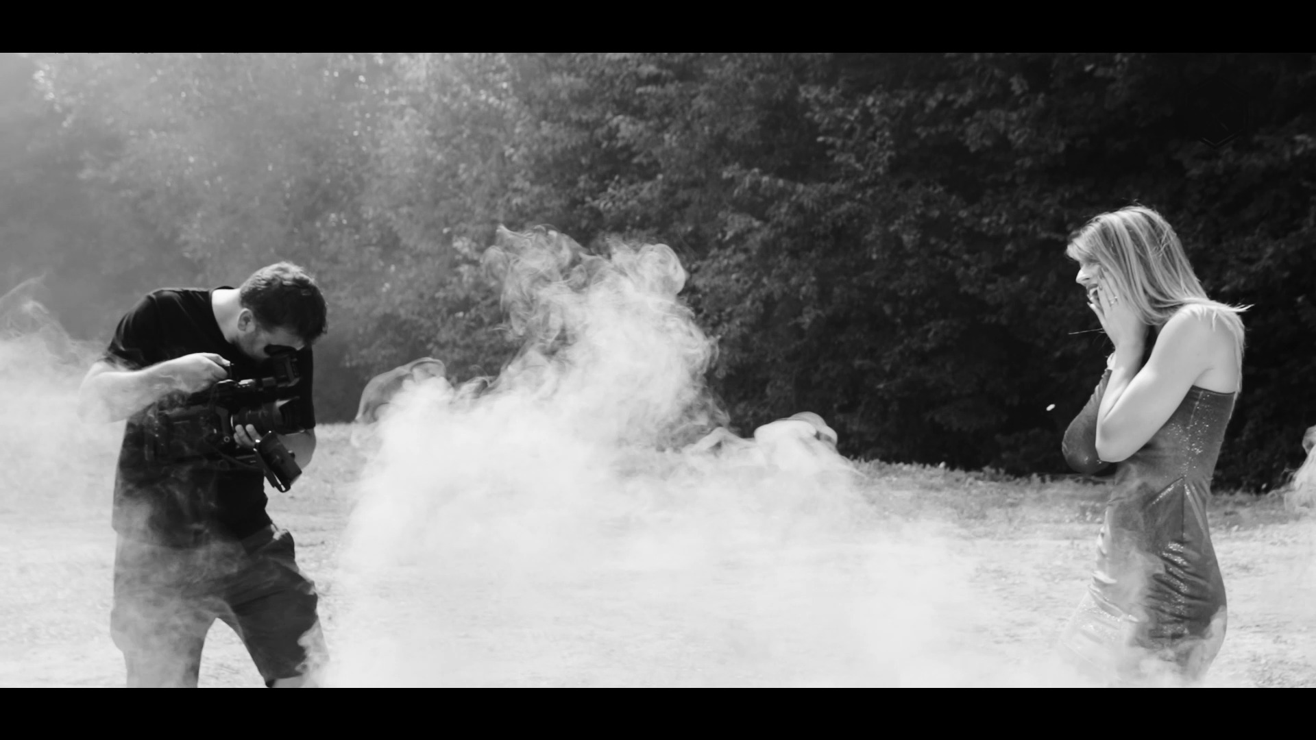 Czarno-białe ujęcie z planu filmowego: kamerzysta z profesjonalnym sprzętem filmuje kobietę w sukience, otoczoną dymem na tle drzew.