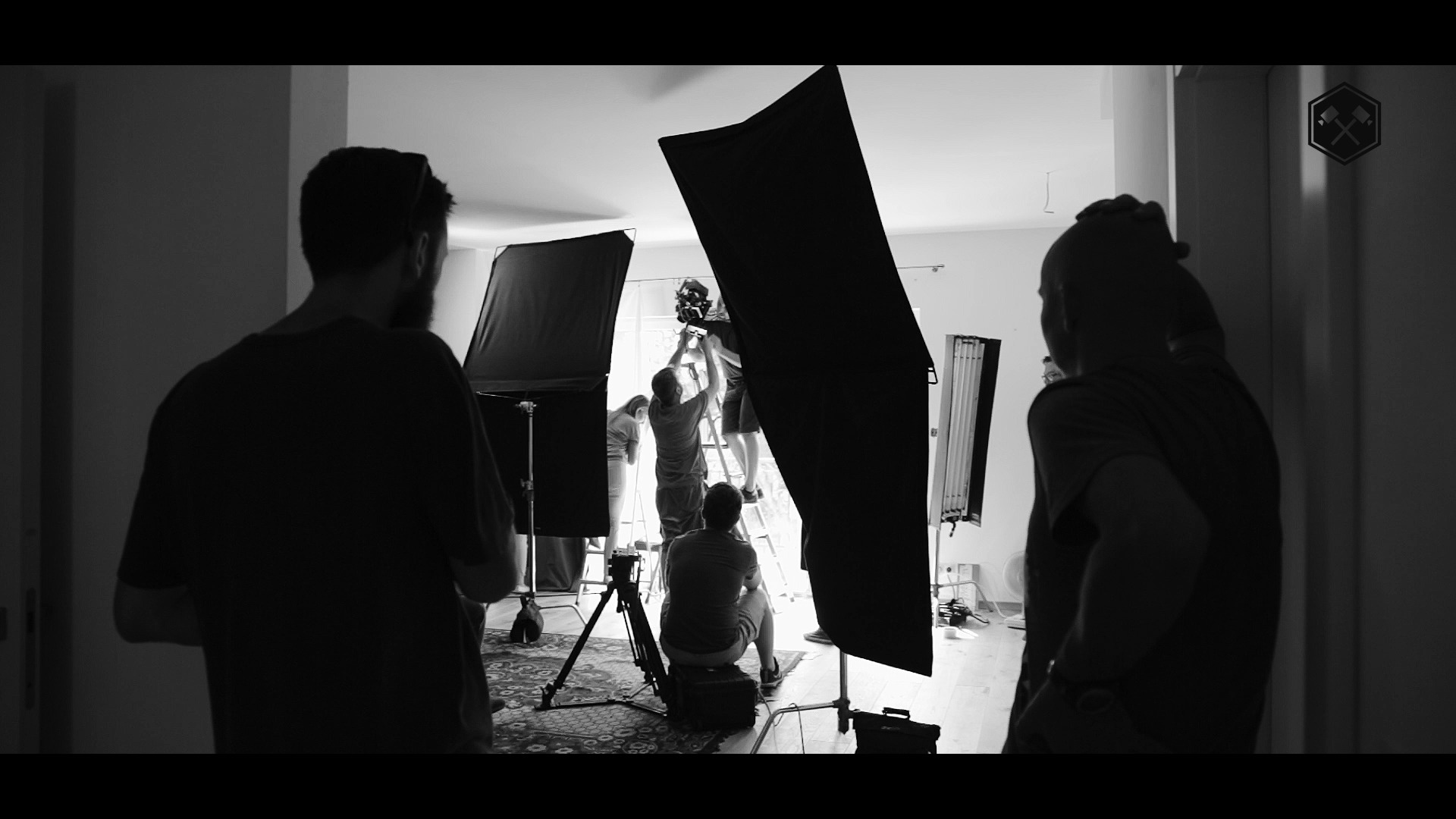 Czarno-białe ujęcie zza kulis: ekipa filmowa ustawia oświetlenie i kamerę w jasnym pomieszczeniu, widoczne softboxy, drabina i osoby w trakcie pracy.