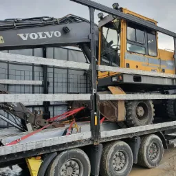 Żółta koparka kołowa Volvo EW200 zabezpieczona czerwonymi pasami transportowymi na niskopodwoziowej platformie ciężarówki, widok z boku, szare niebo